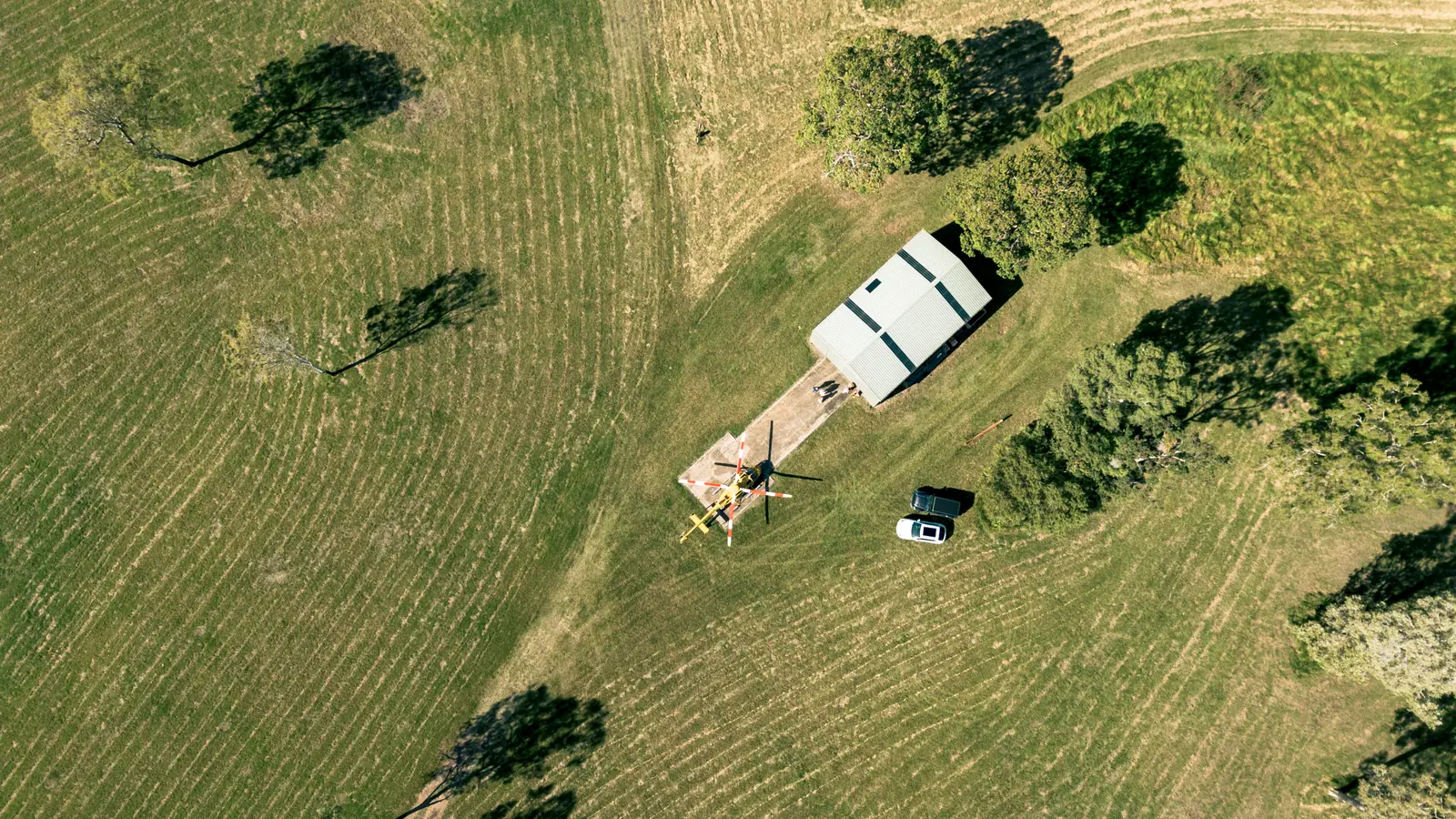 Straight down on the shed and its pad: a yellow helicopter parked with its blades tied, two cars beside it and the grass mown in stripes.